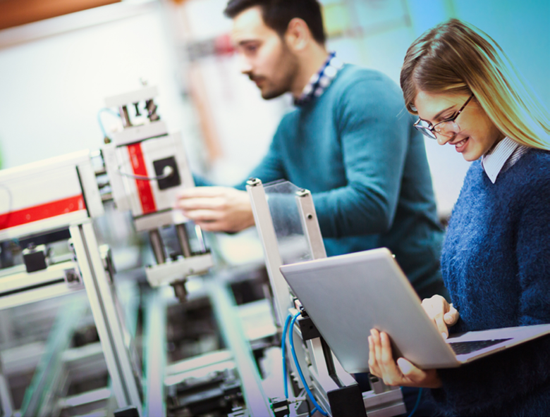 A woman works on a laptop in a manufacturing environment as a man adjusts machinery on an automated production line.