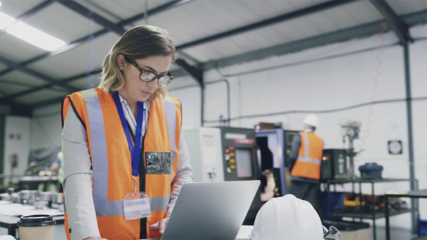 A professional in an industrial workspace reviewing CAD designs on a laptop.