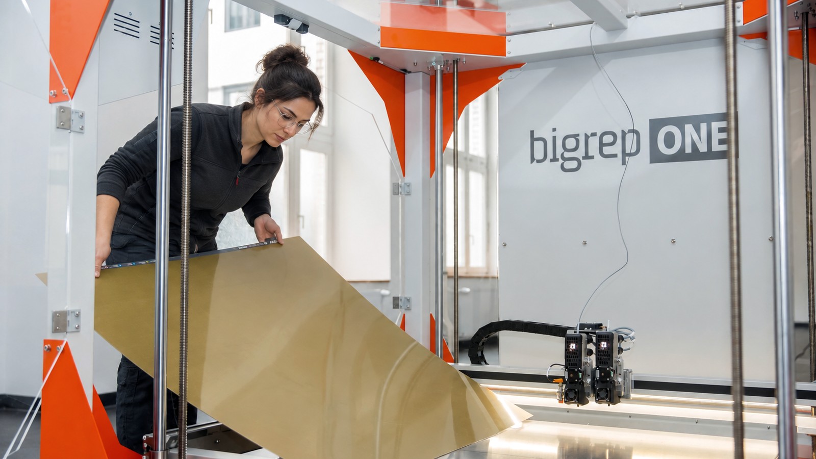 Operator lifting a large gold-coloured printed panel out of a BigRep ONE large format 3D printer, the part roughly the width of her arm span.